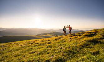 Two people are standing on a green meadow and celebrating the sunset. In the background, there are gentle hills and a clear sky.