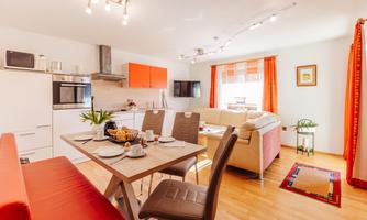 A bright and modern apartment with an open kitchen and a cozy dining area. The windows are adorned with orange curtains, creating an inviting atmosphere.