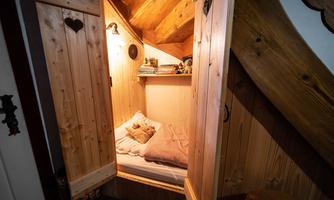 A cozy sleeping spot under a wooden staircase. The room is furnished with a bed and some bookshelves, and the warm wooden tones create an inviting atmosphere.