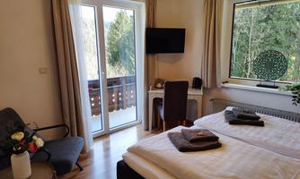 A cozy room with two beds, a table and chair, as well as a television. Large windows offer a view of nature and a balcony.