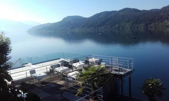 A tranquil lake landscape with gentle hills in the background. On a terrace, there are tables and chairs to enjoy the view.