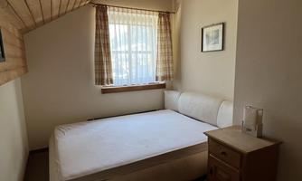 A simple bedroom with a double bed and a bedside table. Large windows with curtains let in plenty of light.