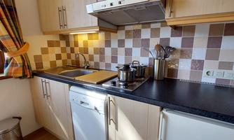 A modern kitchen with wooden cabinets and a black work area. The walls are decorated with brown-beige tiles, and there is a dishwasher as well as cooking utensils.
