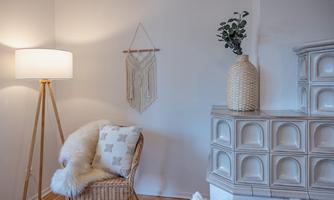 A cozy living room with a wicker chair and a floor lamp. On the wall hangs a macramé, and next to the chair stands a vase with fresh plants.