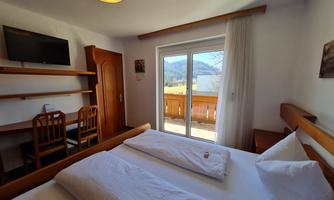 A cozy room with a double bed and two pillows. There is a table, chairs, and a view of the balcony overlooking the mountains.