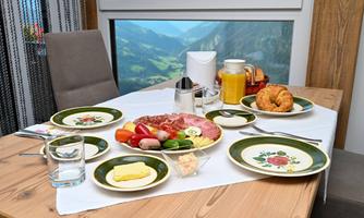 A hearty breakfast on a wooden table featuring various foods. Among them are sausage, vegetables, cheese, croissants, and a drink, with a picturesque view in the background.