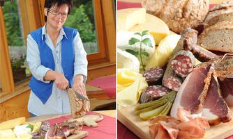 A woman is cutting bread in a cozy environment. On the table are different types of cheese, sausage, and fresh vegetables.