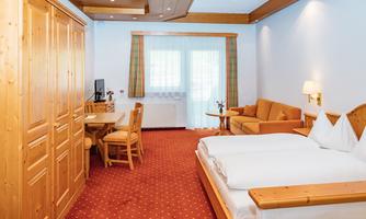 A cozy hotel room with wooden panels and red carpet. It features a double bed, a table with chairs, and a comfortable sitting area.