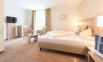 A modern hotel room with a large bed, a television, and comfortable armchairs. Bright colors and natural lighting create an inviting atmosphere.