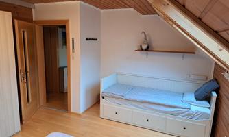 A cozy room with a wooden ceiling and a bed. There is an open door to an adjoining room and a small shelf.