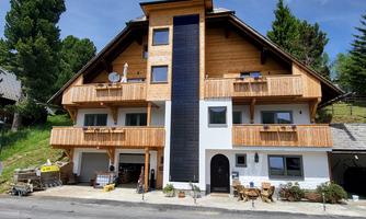 A modern wooden house with a bright entrance and several balconies. It is located in a rural setting surrounded by trees.