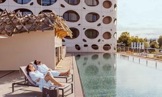 A relaxing pool area with two people in bathrobes resting on loungers. In the background, there is a modern building with round windows.