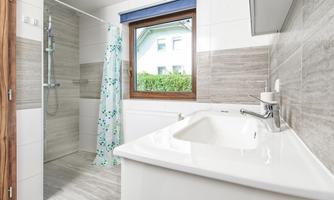 A modern bathroom with a shower and a sink. The bright decor is illuminated by a window that offers a view of greenery.