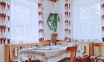 A cozy dining area with wooden chairs and a table, covered with a tablecloth. The windows are adorned with curtains, and a plant hangs on the wall.
