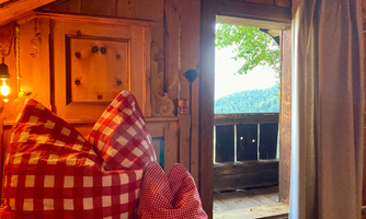 A cozy wooden room with checkered cushions. Through the open window, you can see the green mountains and a clear sky.