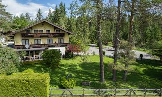 A cozy house surrounded by trees and a green lawn. The sky is clear and the surroundings are calm and idyllic.
