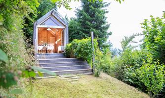A modernly designed wooden house stands on a meadow, surrounded by trees. The steps lead to the open entrance area of the house.