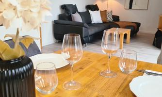 A modernly furnished room with a dining area and a cozy living room. There are glasses and dishes on the table, while a stylish decoration can be seen in the background.