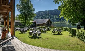 A quiet meadow by the water with several tables and chairs. In the background, trees and a hill can be seen.