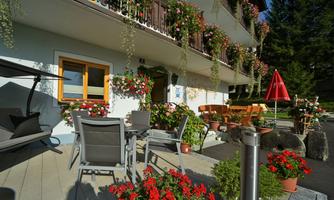 A cozy outdoor area with blooming flowers and terrace furniture. In the background, a balcony railing with many plants can be seen.