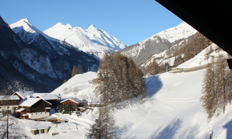 A picturesque mountain landscape with snow-capped peaks and small chalets. The sky is clear and blue, creating a tranquil winter atmosphere.
