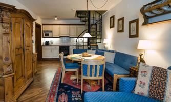 A cozy living room with blue sofas and a round dining table. In the background, you can see a small kitchen and a spiral staircase.