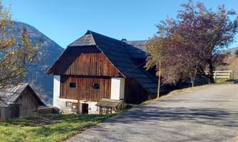 A charming, traditional farmhouse stands on a quiet road. In the background, gentle mountains and a few trees can be seen.