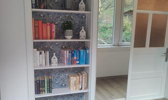 A modern room with a bookshelf displaying colorful books and small decorative elements. Light floods the room through large windows, creating a bright atmosphere.
