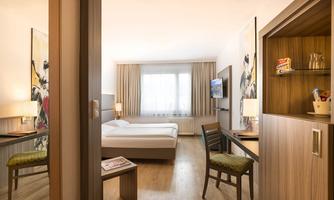 A modern hotel room with a double bed, desk, and television. Large windows provide plenty of daylight.
