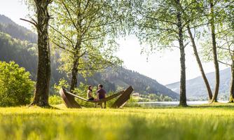 A quiet garden by the water with green meadows and tall trees. Two people are comfortably sitting in a modern wooden lounge chair, enjoying nature.