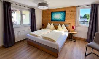 A bright, modern bedroom with a large double bed and beige pillows. The walls are partly made of wood and the windows provide plenty of natural light.