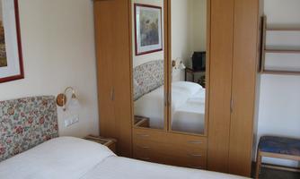 A simple bedroom with a bed, a wardrobe, and a chair. Pictures are hanging on the wall and there is a bright, friendly atmosphere.