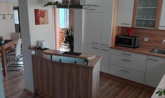 A modern kitchen with a wood finish and an open layout. In the background, plants and a dining area are visible.