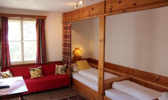 A cozy room with wood paneling and two single beds. The room has large windows with curtains and a red couch.