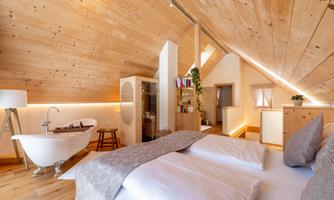 A cozy attic room with wooden walls and a large window. The room is stylishly furnished and features a freestanding bathtub and a comfortable bed.