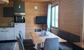 A modern dining room in a wooden apartment with a large table and comfortable chairs. In the background, a kitchenette and a window with natural light are visible.