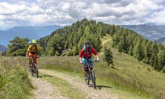 Two cyclists are riding on a gravel path through a green landscape. In the background, gentle hills and a cloudy sky can be seen.