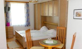 A cozy bedroom with a double bed and a small dining table. The room has large windows that let in plenty of daylight.