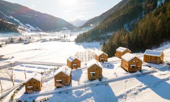 A picturesque winter village with modern wooden chalets, surrounded by snow-covered fields and mountains. The sun is shining brightly, giving the landscape a warm light.