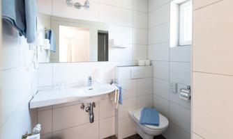 A modern bathroom with white tiles and a sink. It includes a toilet and large windows for natural light.