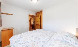 A bright bedroom with two single beds and floral bedcovers. The walls are mostly white, and there is a wooden wardrobe as well as windows with curtains.