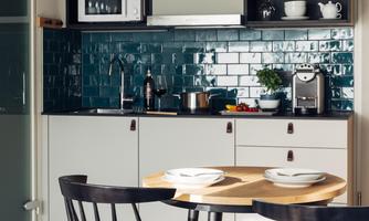 A modern kitchen with blue-green tiles and a minimalist setup. A round wooden table with two plates and chairs invites you to dine.