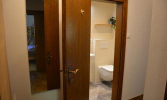 A hallway with a wooden door and a mirror. In the background, a bathroom with a toilet and a small plant can be seen.