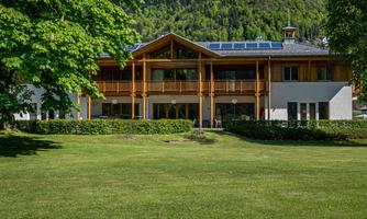 A modern building with wood cladding and a spacious balcony.  
Surrounded by green grass and trees in a mountainous landscape.