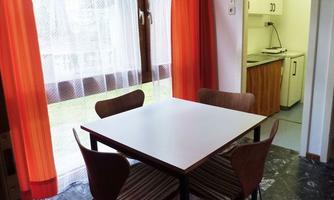 A small dining area with a white table and four chairs. In the background, a kitchen is visible, and the room has large windows with red curtains.