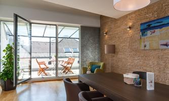 A modern room with a large window that overlooks a terrace with wooden chairs and a table. The walls are designed with natural stone and there are plants that create an inviting atmosphere.