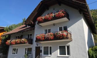 A modern house with multiple balconies, adorned with colorful flowers. The surroundings are green and sunny.