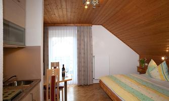 A cozy room with a wooden ceiling and a double bed. There is a small dining area and windows with curtains that let in natural light.