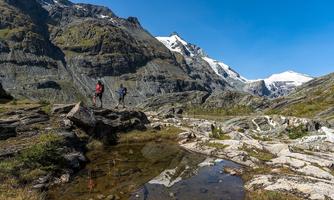Two hikers explore an impressive mountain landscape with snow-capped peaks. A clear sky and a small body of water enhance the beautiful scenery.