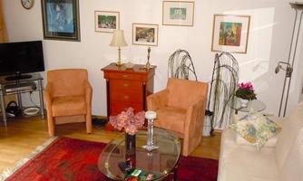 A cozy living room with a red carpet, comfortable armchairs, and a coffee table. Various pictures hang on the walls, and a floor lamp provides lighting.